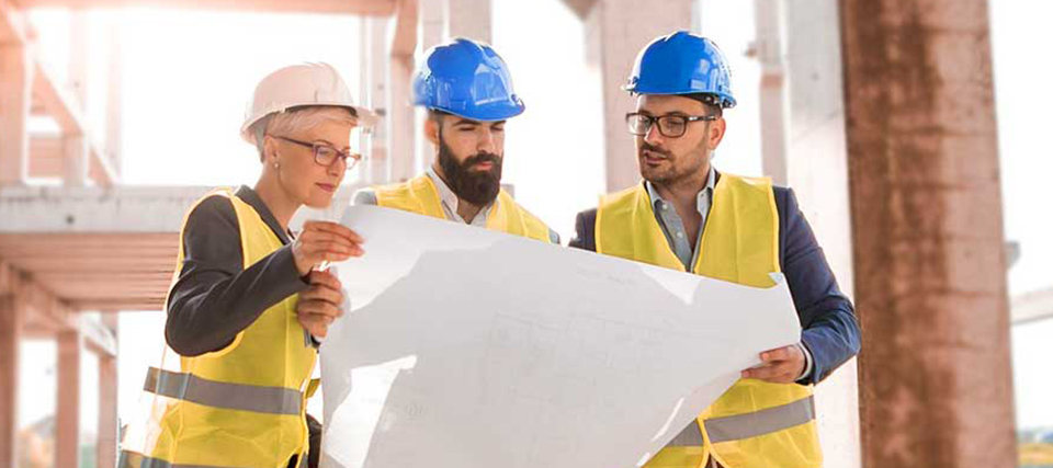 Drei Bauingenieure mit Schutzhelmen und Warnwesten besprechen Baupläne auf einer Baustelle.