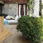 Lifestyle-Ansicht einer Terrasse mit Keramik-Bodenplatten in heller Holzoptik (Farbe Honey), kombiniert mit einer Natursteinwand, Lounge-Möbeln und Grünpflanzen. Im Vordergrund sind drei einzelne Produktmuster der Fliese abgebildet.