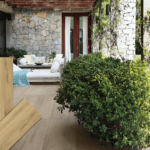 Terrasse mit Keramik-Bodenplatten in authentischer, heller Holzoptik (Farbe Natural), kombiniert mit einer Steinwand und Gartenmöbeln. Im Vordergrund drei Produktmuster der Serie HAT-88.